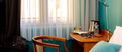 Quarto de hotel com uma cadeira de madeira, uma mesa com luminária, uma cama e uma janela com cortinas leves.