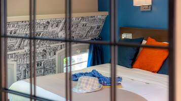 A hotel room with a bed, a blue and orange pillow, a hat, and a wall with a black and white cityscape mural.