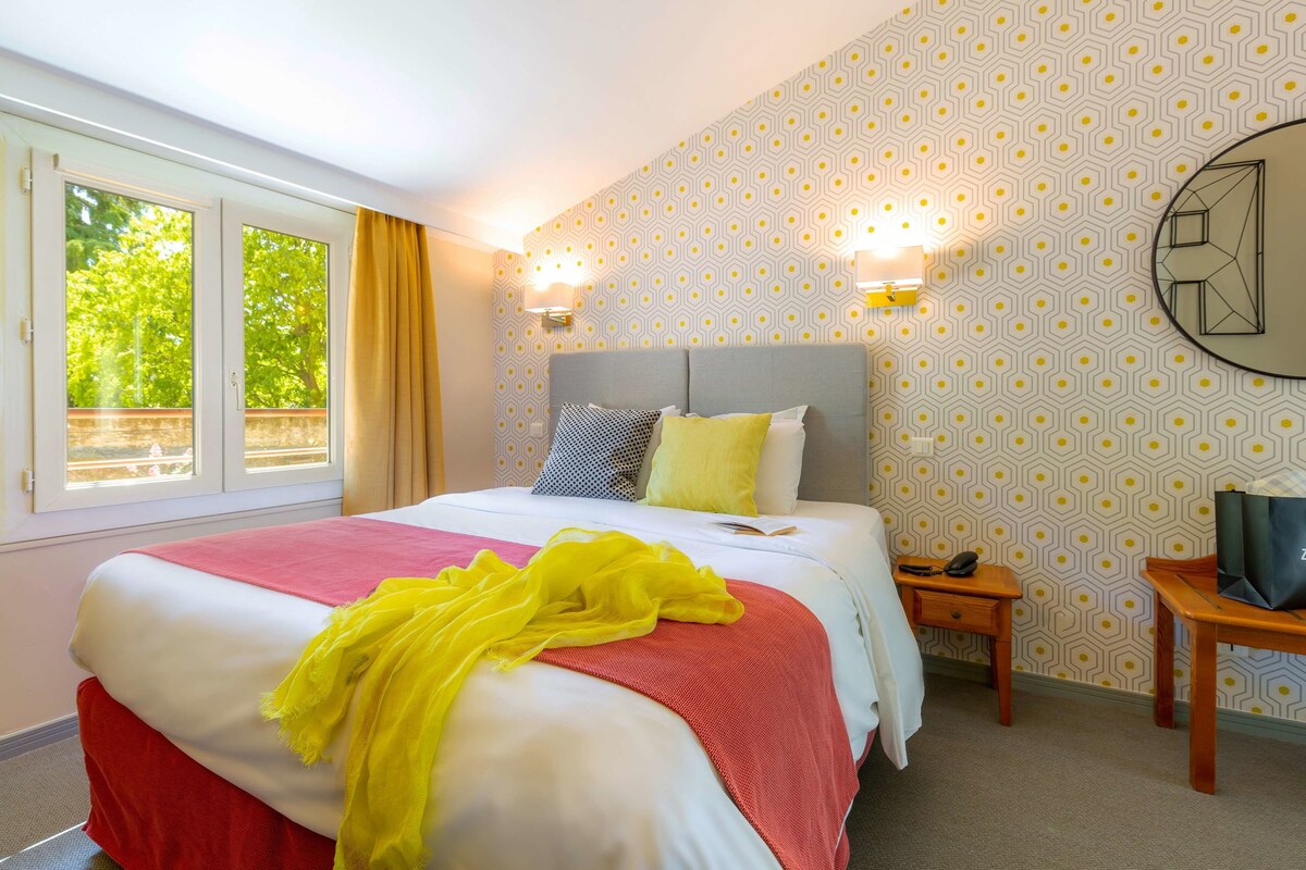 A hotel room with a bed, a nightstand, a chair, a window with curtains, and patterned wallpaper.