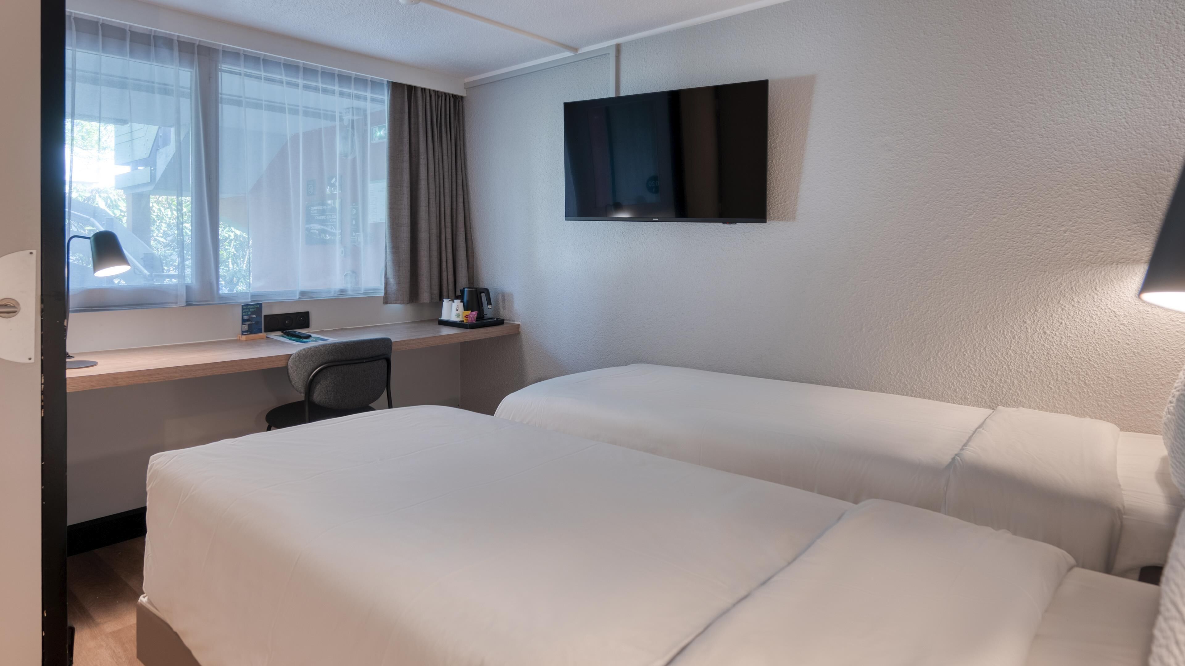 A hotel room with two beds, a desk, a chair, a TV, and a window with curtains.