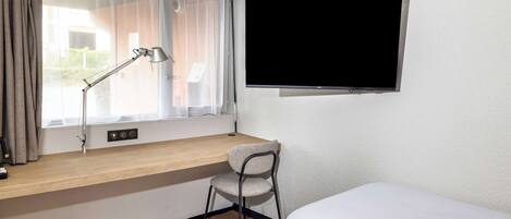A hotel room with a bed, a desk with a chair, a TV mounted on the wall, and a window with curtains.