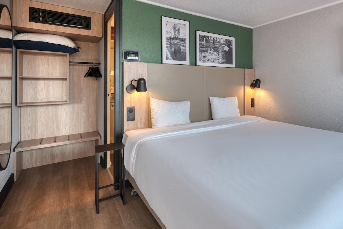 A hotel room with a bed, a wooden wardrobe, a mirror, and two wall-mounted pictures.