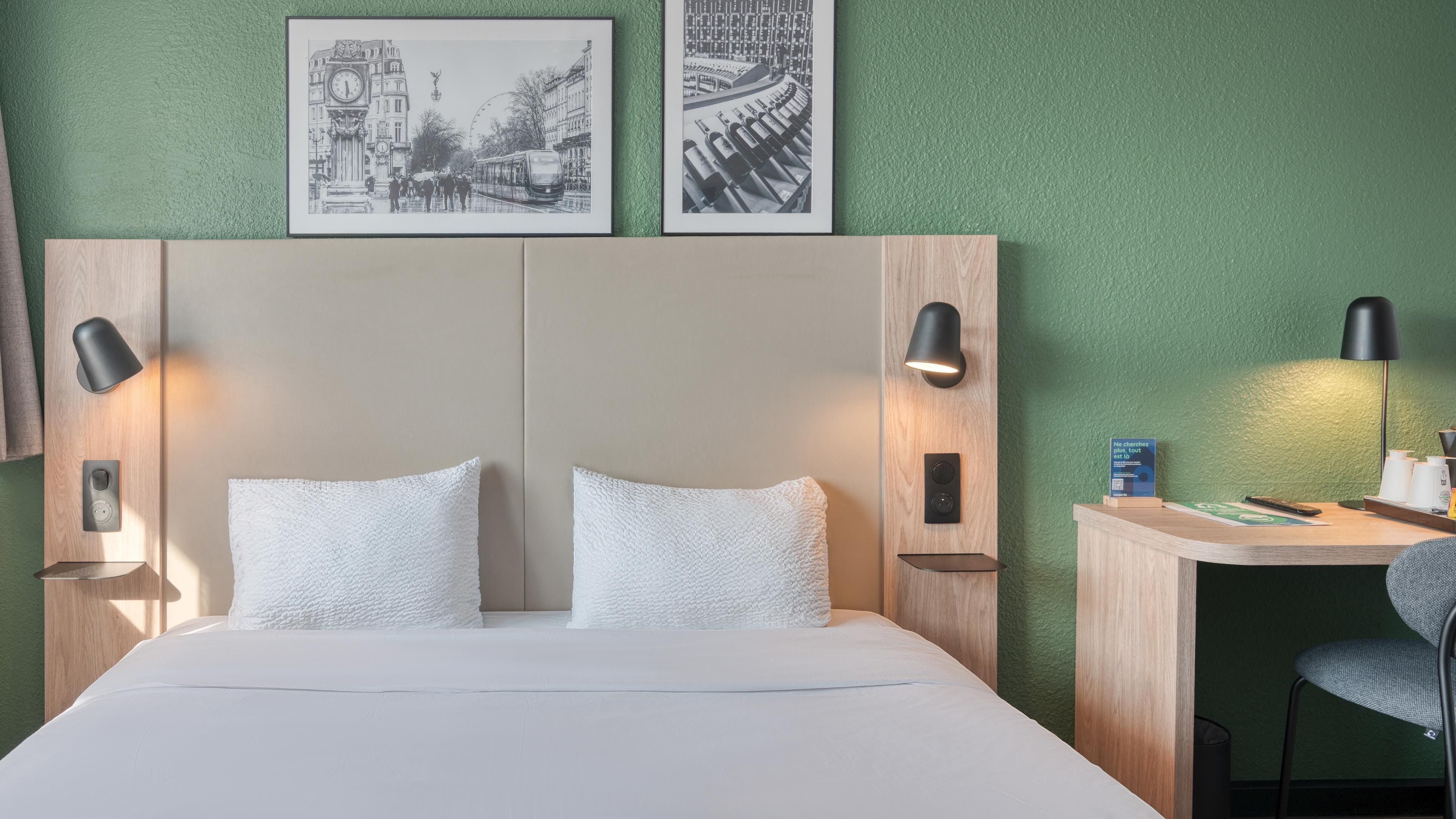 A hotel room with a bed, two pillows, a desk with a chair, a lamp, and two wall-mounted lamps.