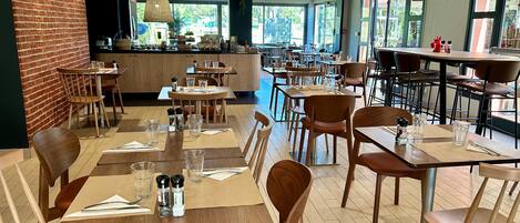 Un restaurant spacieux avec des tables et des chaises en bois, un mur en briques et de grandes fenêtres laissant entrer la lumière naturelle.