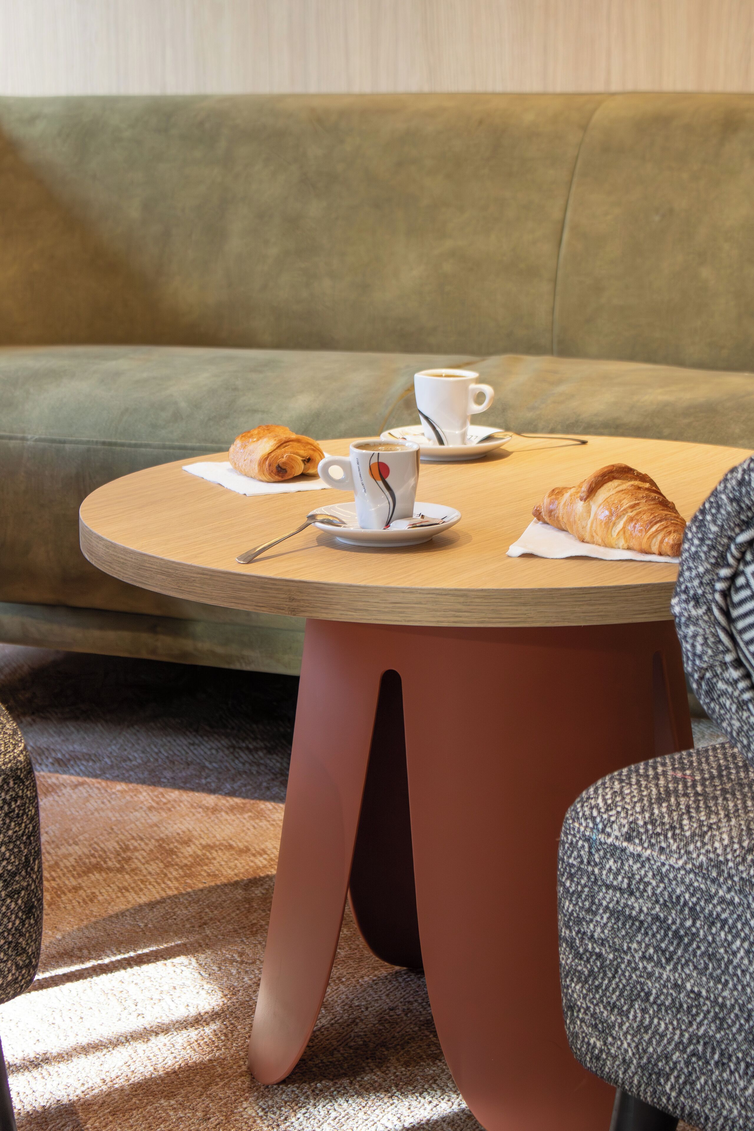 A round table with a croissant and a cup of coffee, a sofa in the background, and a patterned chair in the foreground.