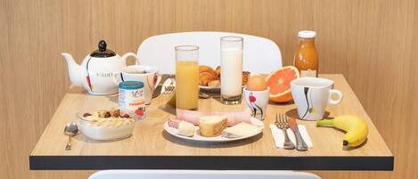 A breakfast table with a teapot, cups, a bottle, a banana, a croissant, and a plate with food.
