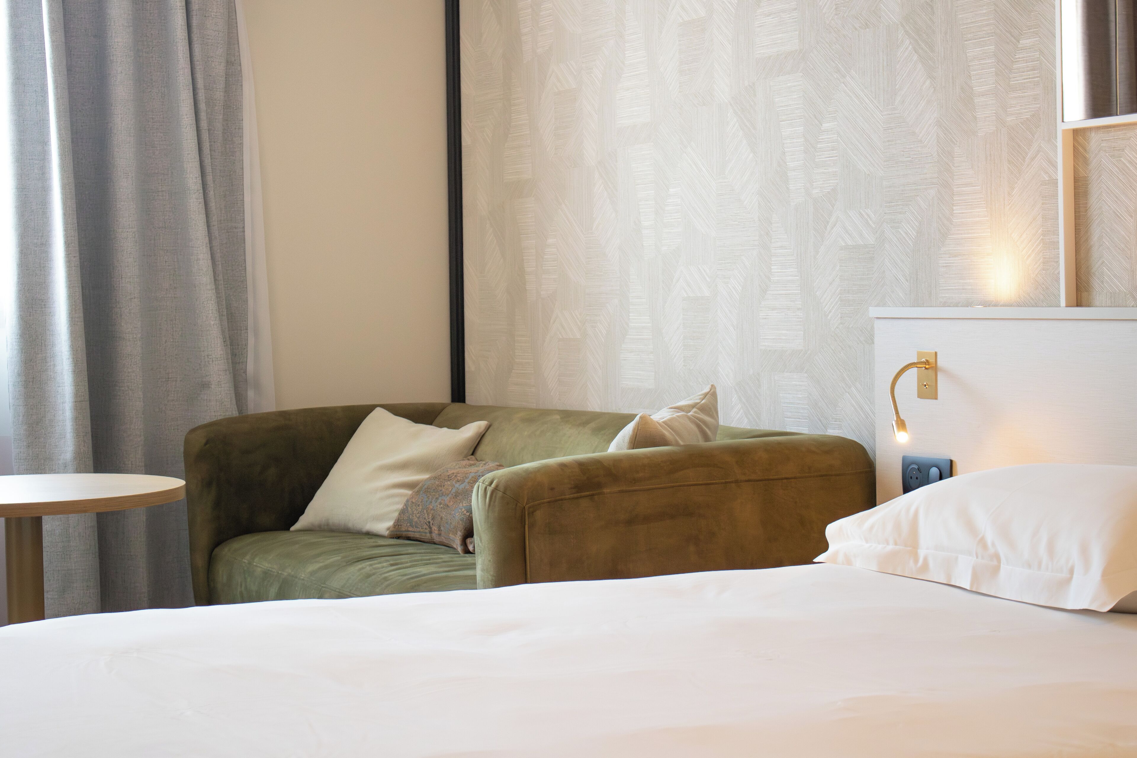 Una habitación de hotel con una cama, un sillón verde, una mesita redonda y una lámpara fijada a la pared.