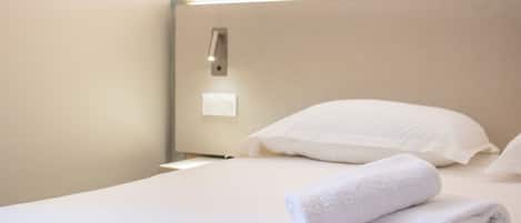 A neatly made bed with white linens and a rolled white towel, a wall-mounted light fixture, and a plain wall.
