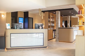 A modern hotel reception area with a digital screen, a self-service beverage station, and a bar with shelves of liquor. - Brit Hotel Avignon Sud (Avignon)
