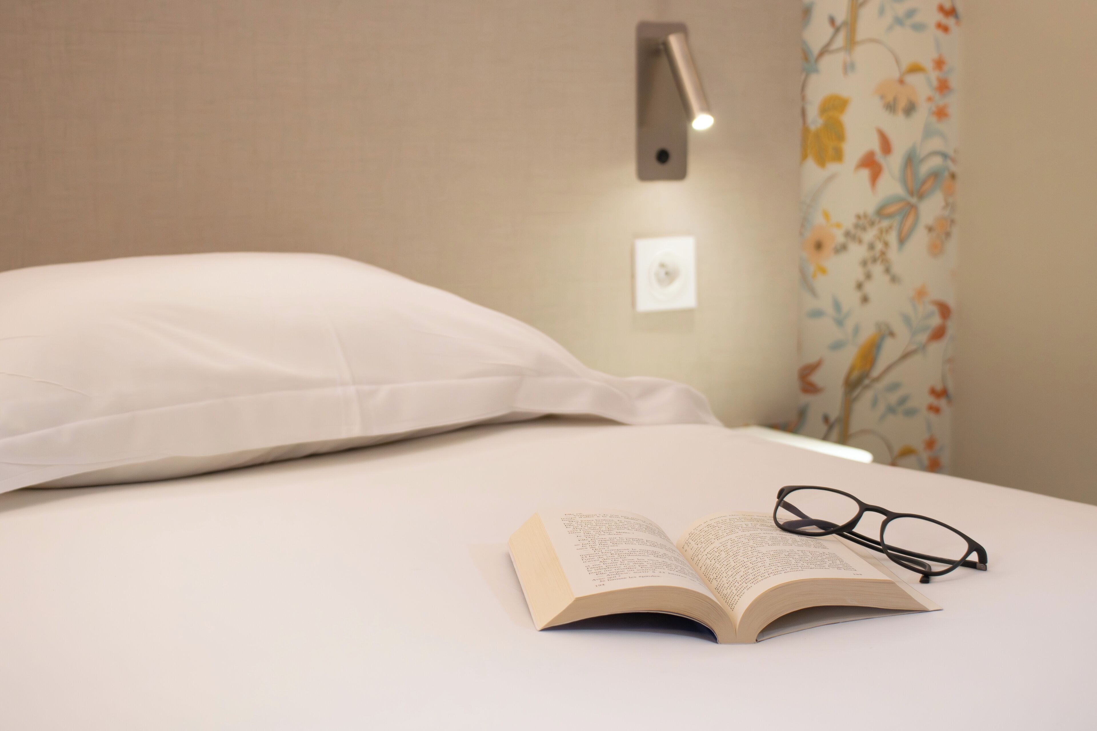 A neatly made bed with a pillow, an open book, and a pair of glasses on the bed.