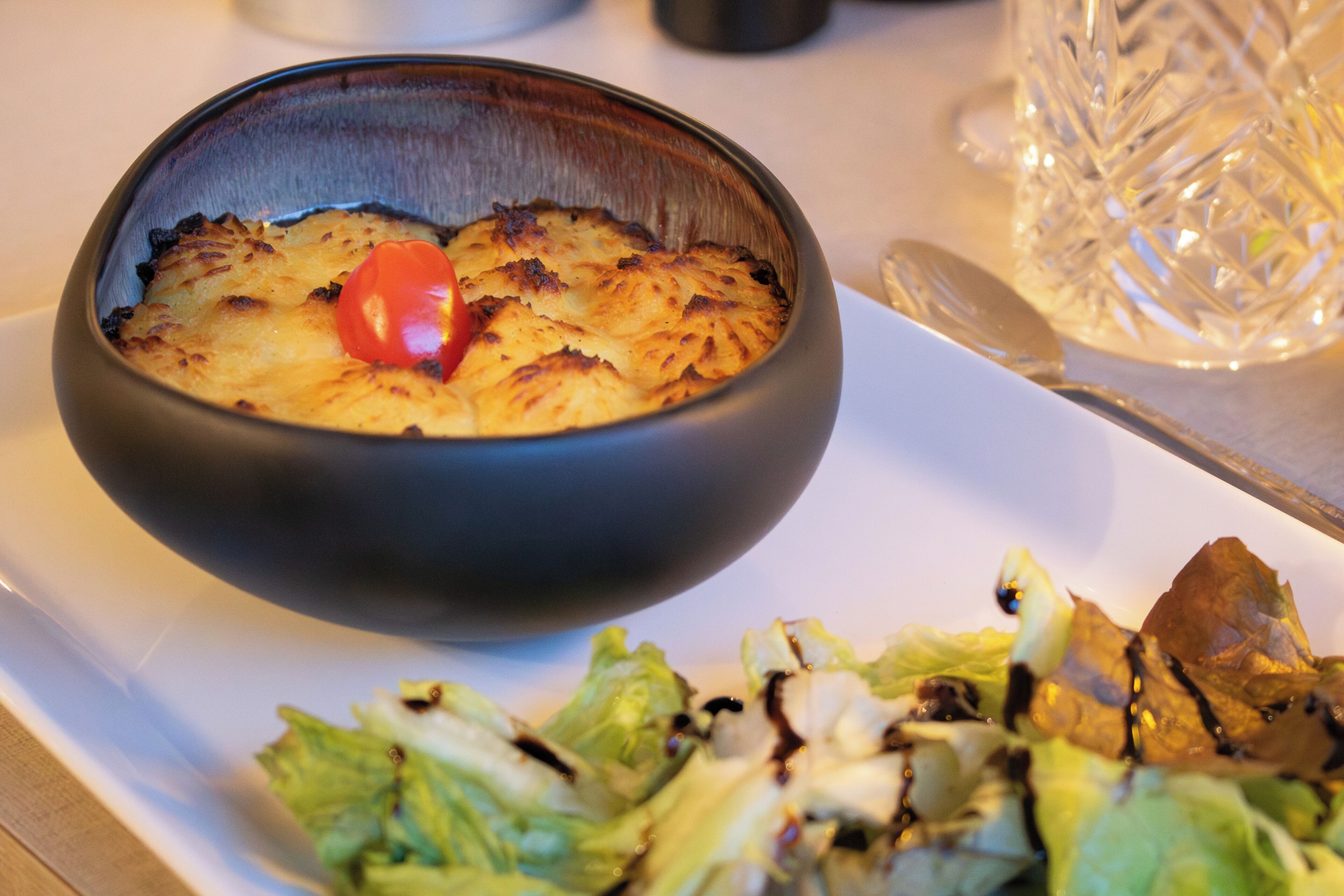 A bowl of food with a cherry on top, a salad with dressing, and a glass on a table.