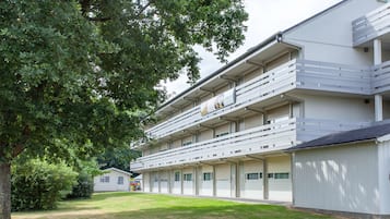 Een hotel met meerdere verdiepingen, een plat dak, diverse balkons en een grote boom op de voorgrond.