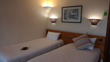 Habitación de hotel con dos camas individuales, cada una con una almohada y un libro. Hay luces fijadas a la pared y un cuadro enmarcado sobre las camas.