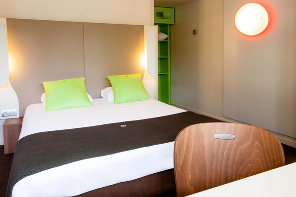 A hotel room with a bed, green pillows, a wooden chair, and a wall-mounted light. - Hotel Campanile Angers Ouest - Beaucouzé (Beaucouze)