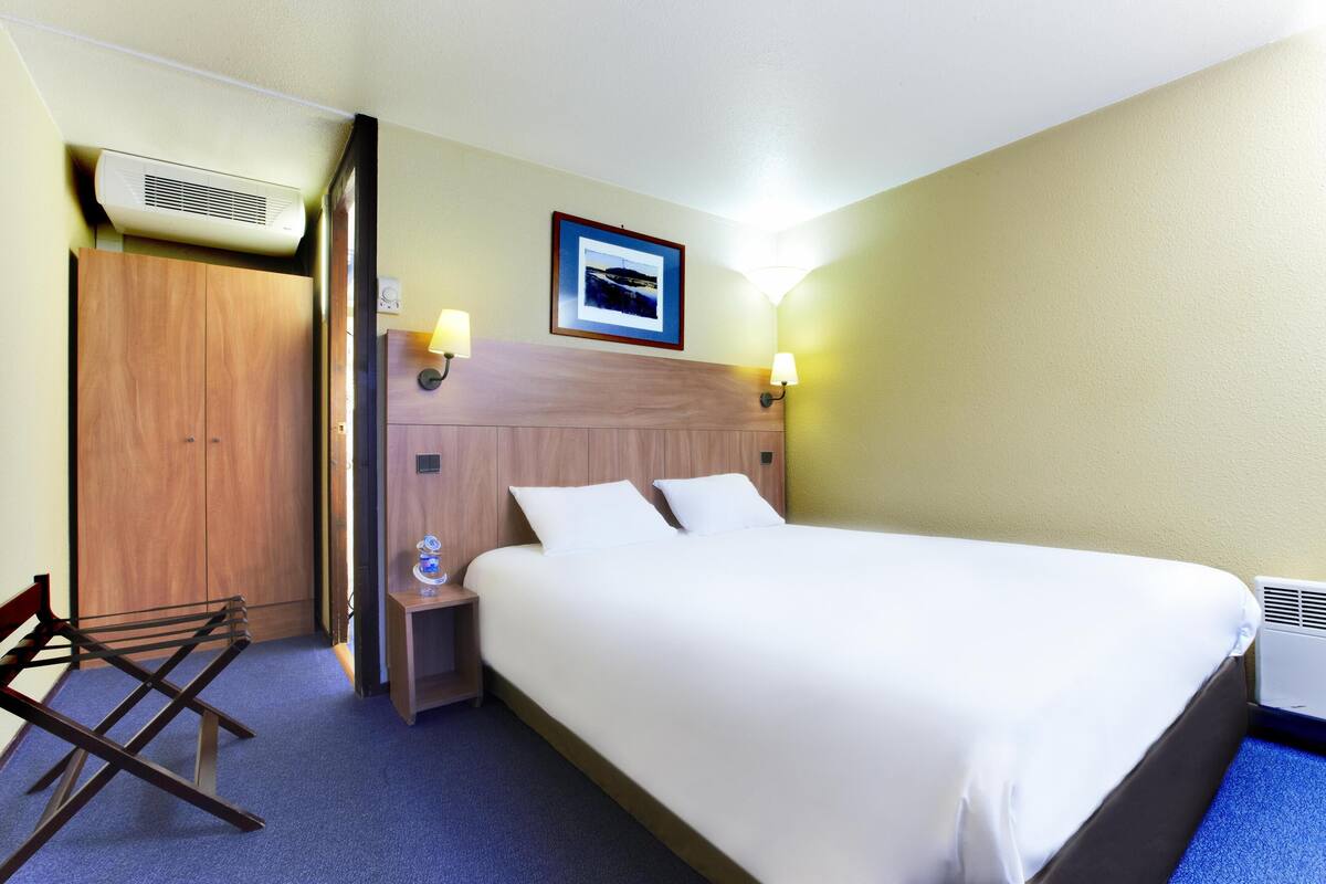A hotel room with a bed, a wooden wardrobe, a nightstand with a lamp, and an air conditioning unit.