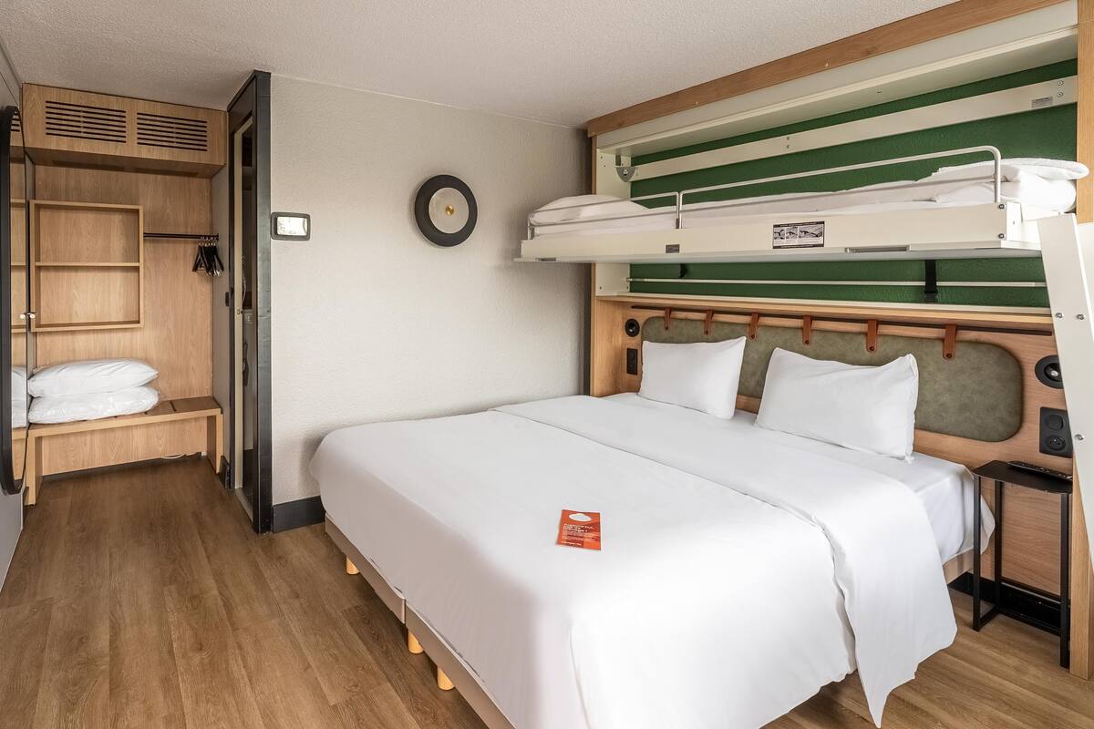 A compact room with a bunk bed, a wooden wardrobe, a bench, and a small bedside table.