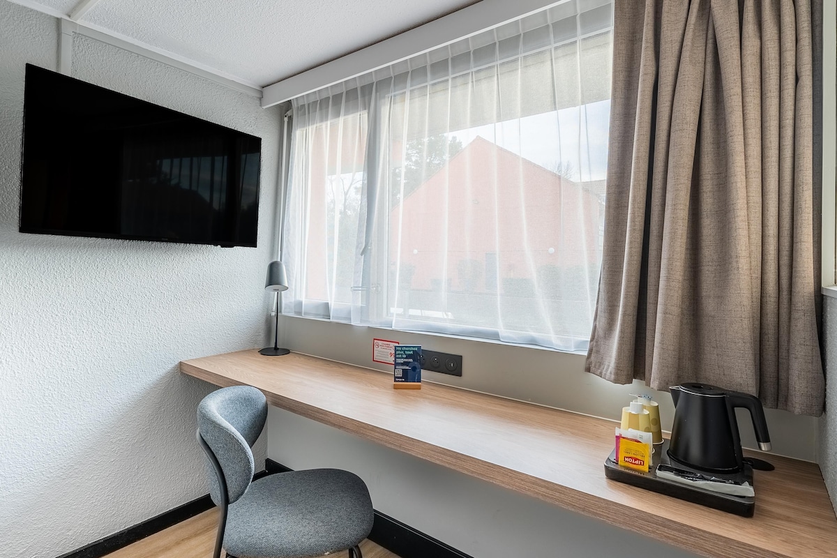 Una habitación pequeña y moderna con un escritorio de madera, una silla, un televisor montado en la pared, una ventana con cortinas translúcidas y una cafetera sobre el escritorio.