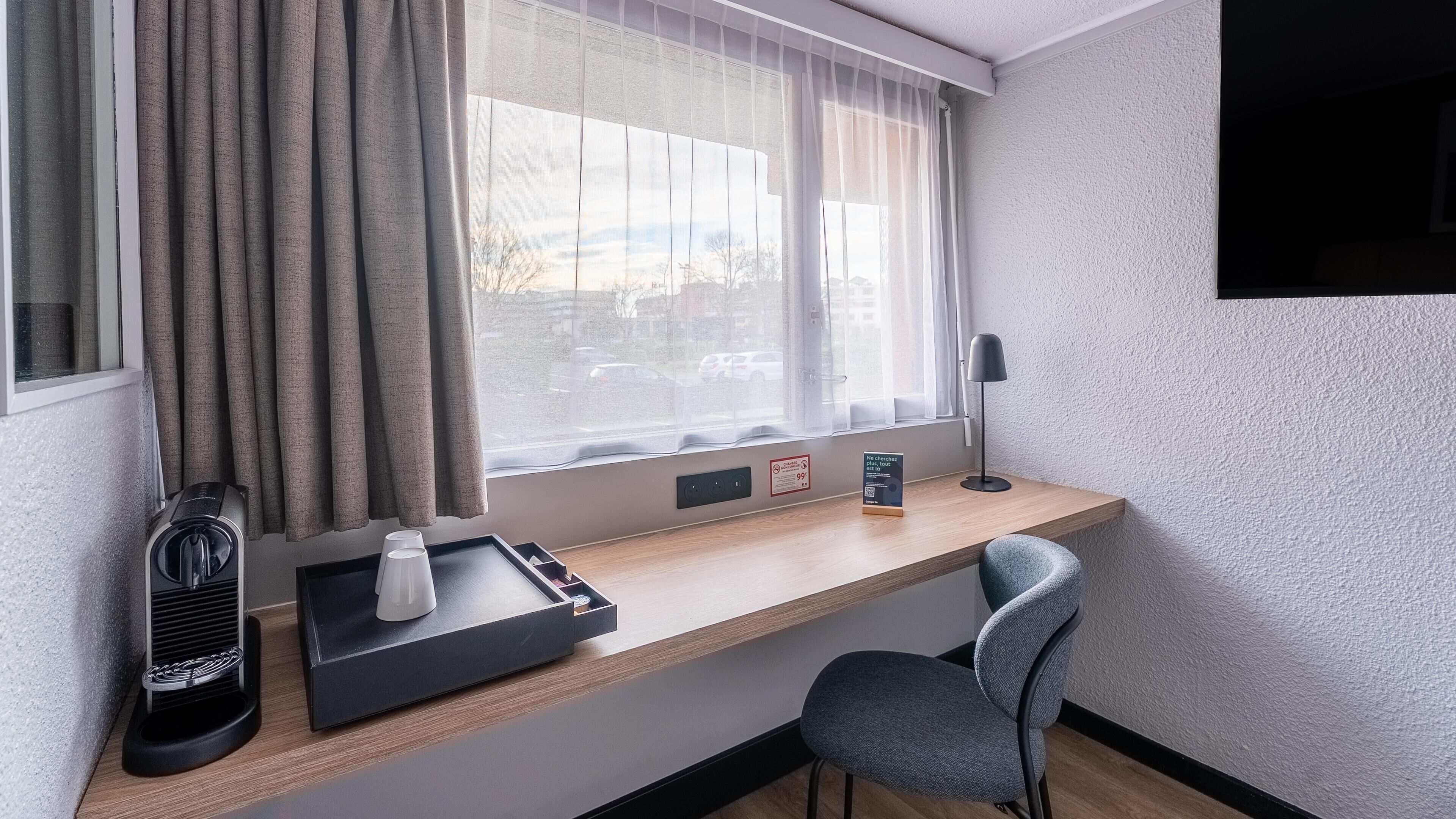 A hotel room with a desk, a chair, a window with curtains, a lamp, and a tray with a coffee maker and cups.