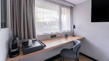 A hotel room with a desk, a chair, a window with curtains, a lamp, and a tray with a coffee maker and cups.