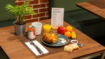 Una mesa de desayuno con un menú, una planta, un vaso de jugo de naranja, frutas, croissants y un frasco pequeño de mermelada.