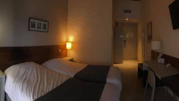 A hotel room with two beds, bedside lamps, a desk with a chair, a television, and a door leading to another room.