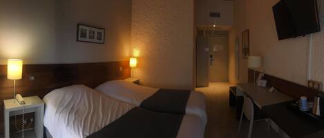 Une chambre d’hôtel avec deux lits, des lampes de chevet, un bureau avec une chaise, une télévision et une porte donnant sur une autre pièce.