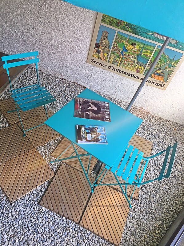 A small outdoor seating area with a blue table and matching chairs, surrounded by pebbles and wooden flooring.