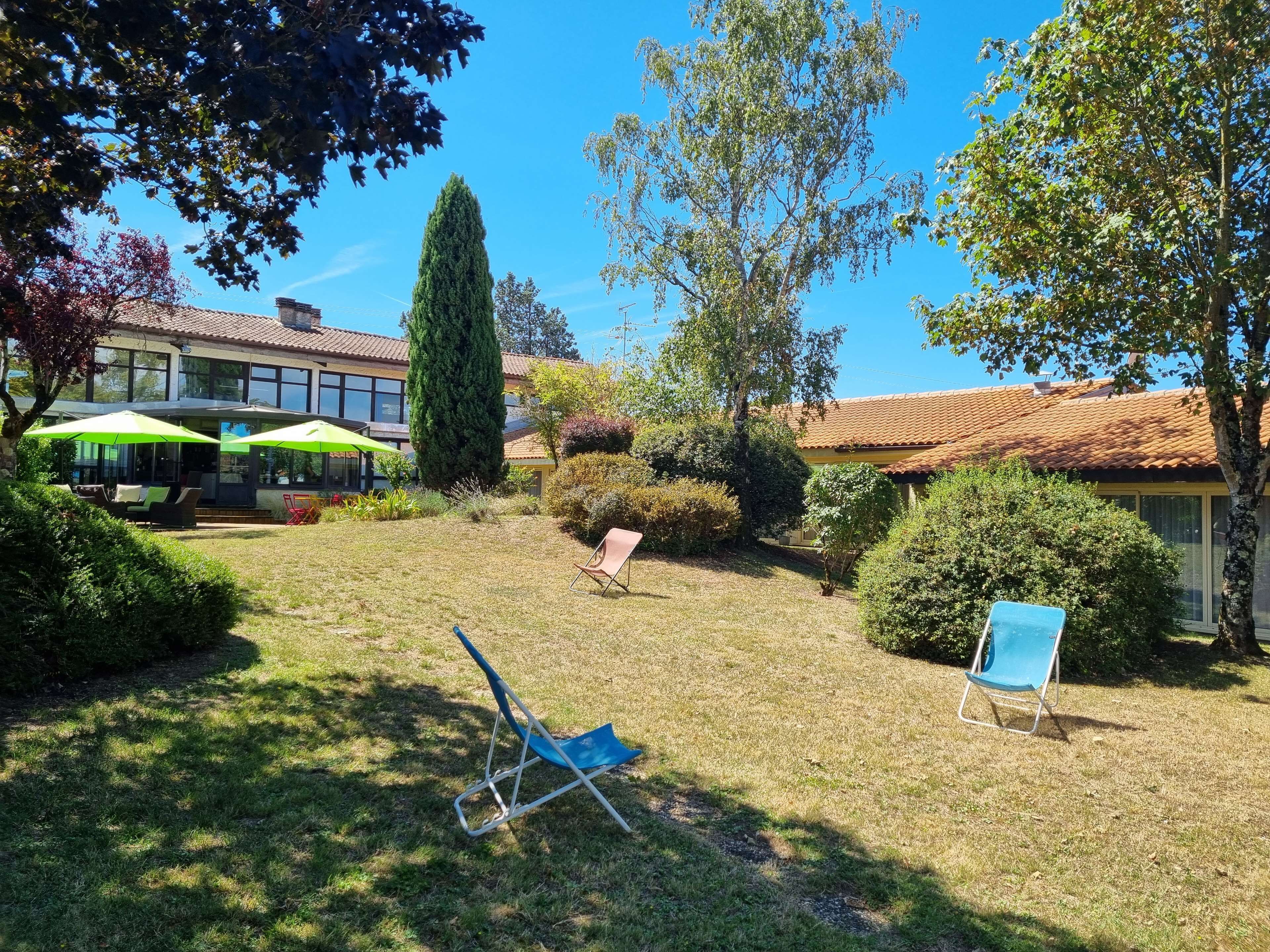A sunny courtyard with a lawn, two lounge chairs, a tall tree, and a building with a tiled roof.