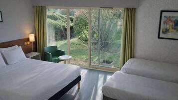 A hotel room with two beds, a small table, a chair, a lamp, and a large window with curtains.