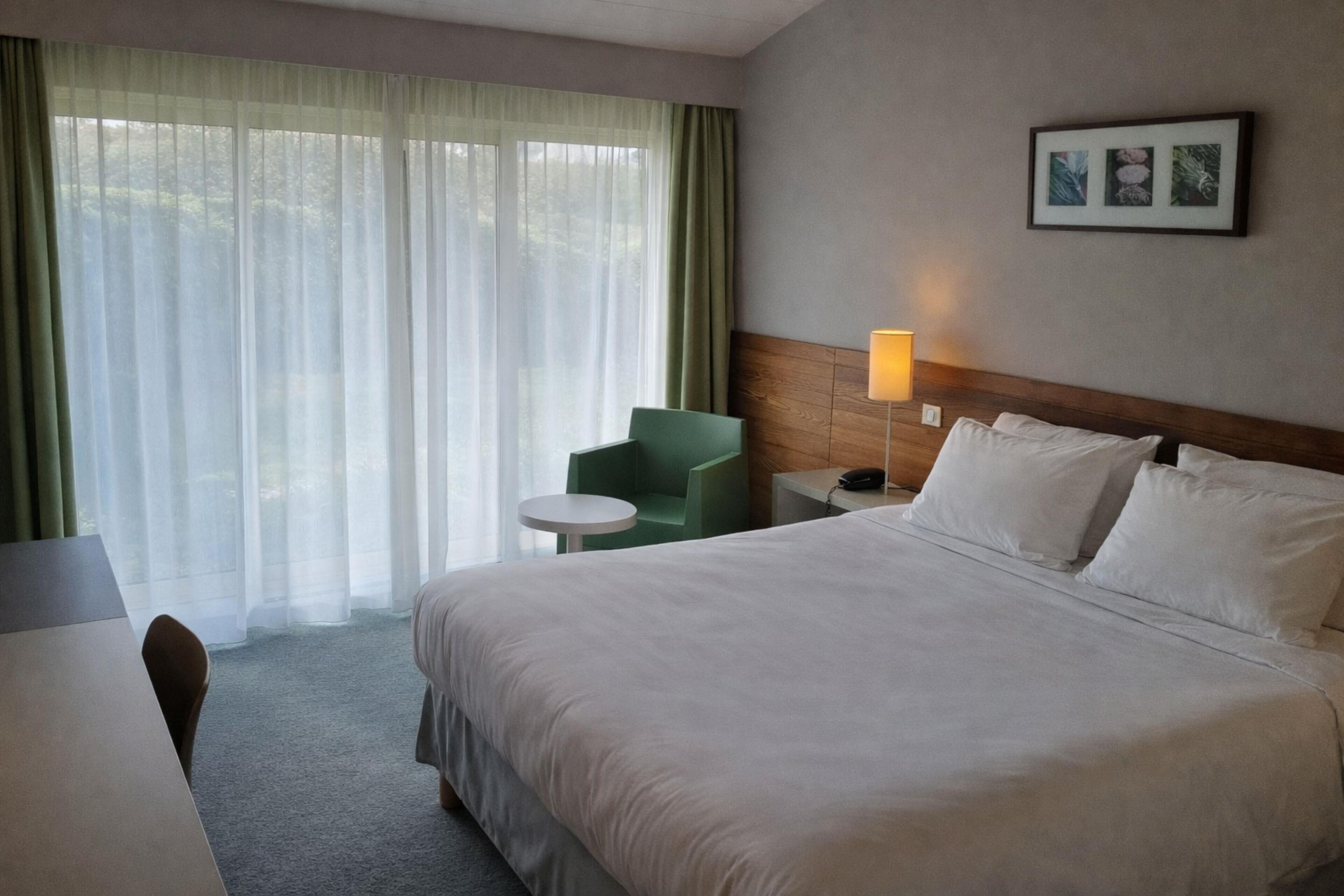 A hotel room with a bed, a desk, a chair, a lamp, and a window with curtains.