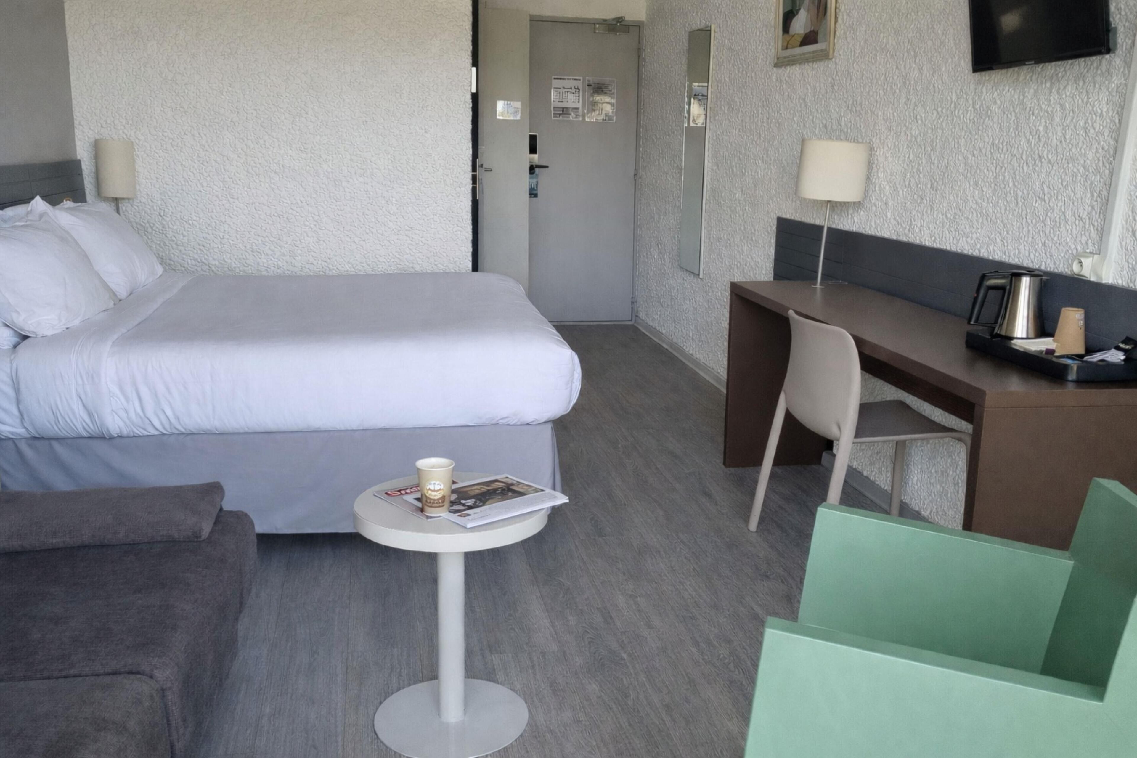 Ein Hotelzimmer mit einem Bett, einem Schreibtisch, einem Stuhl, einem kleinen Tisch mit Zeitschrift und Tasse sowie einem an der Wand befestigten Fernseher.