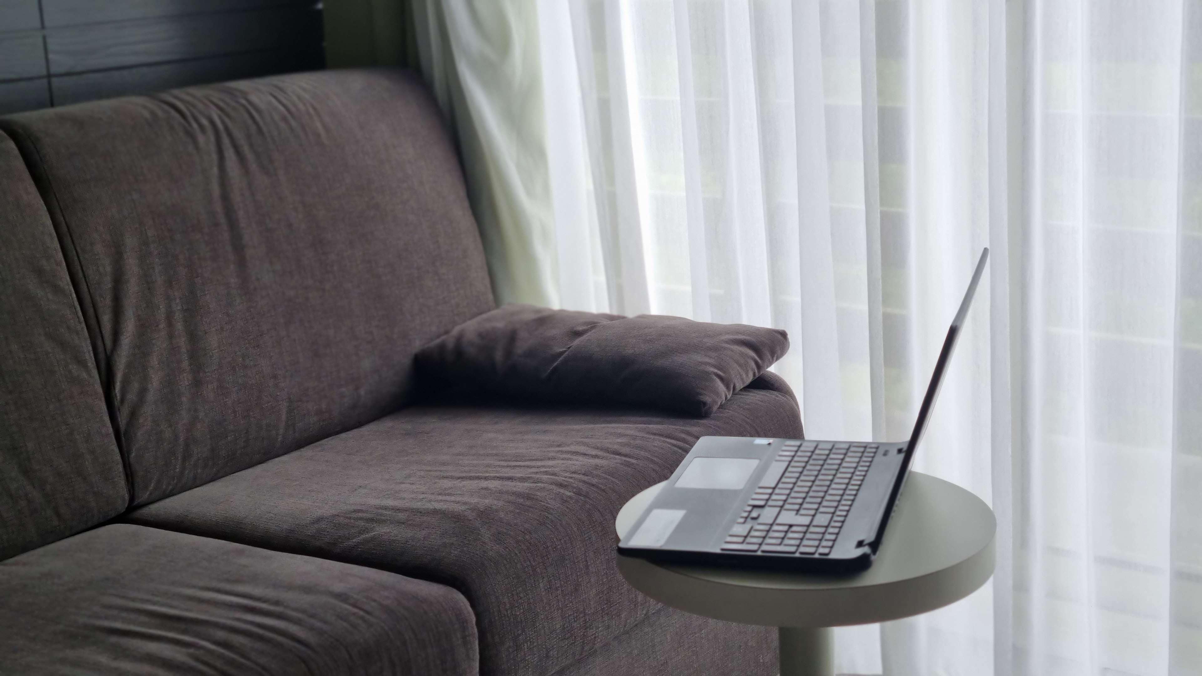 Una computadora portátil sobre una mesa redonda pequeña, junto a un sofá gris con un cojín.