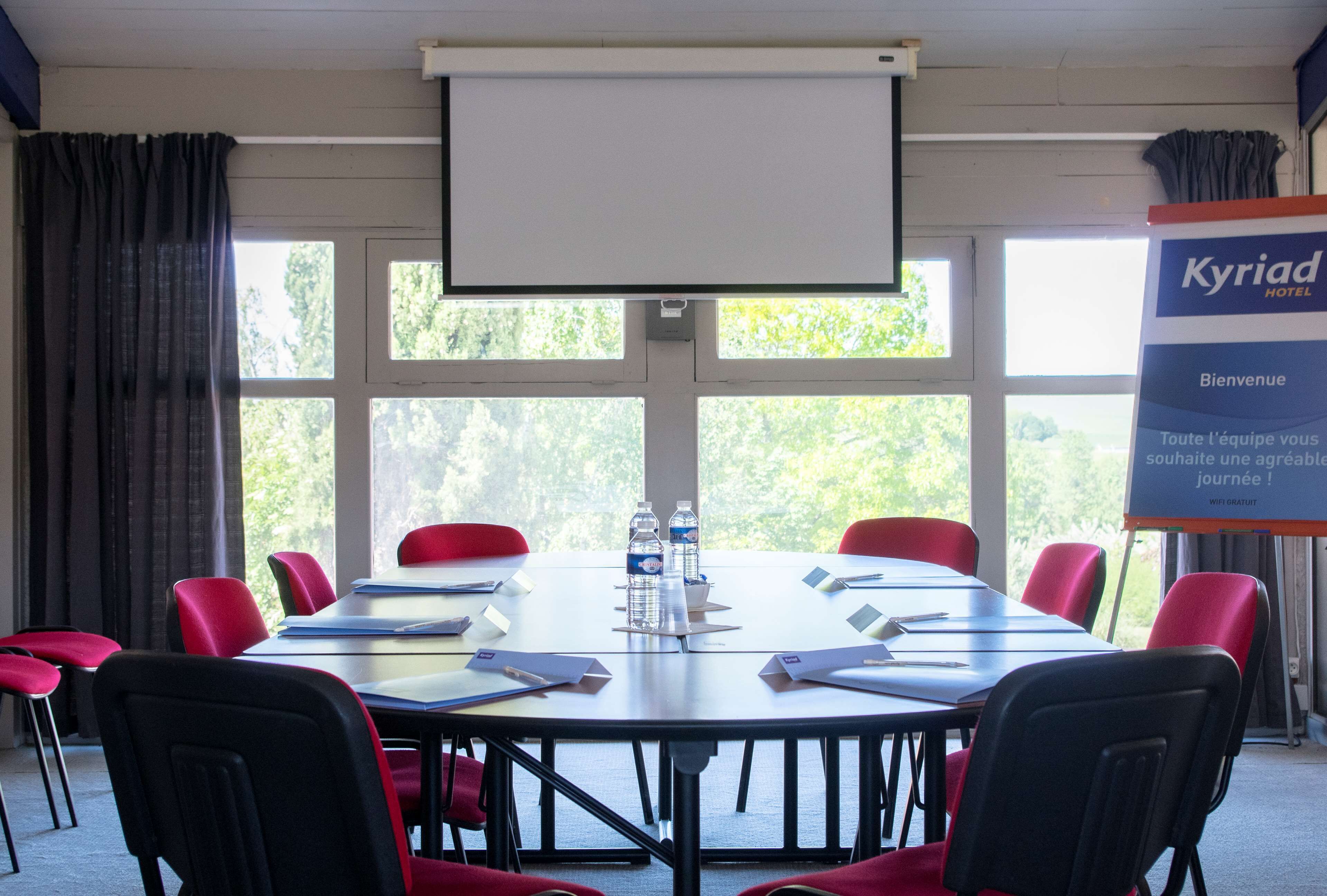 Ein Konferenzraum mit großem Tisch, roten Stühlen, einer Projektorleinwand und einem Banner für Kyriad Hotel.