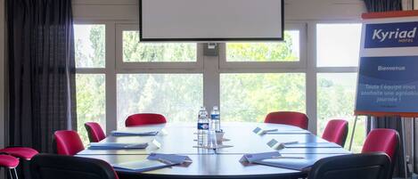 A conference room with a large table, red chairs, a projector screen, and a banner for Kyriad Hotel.