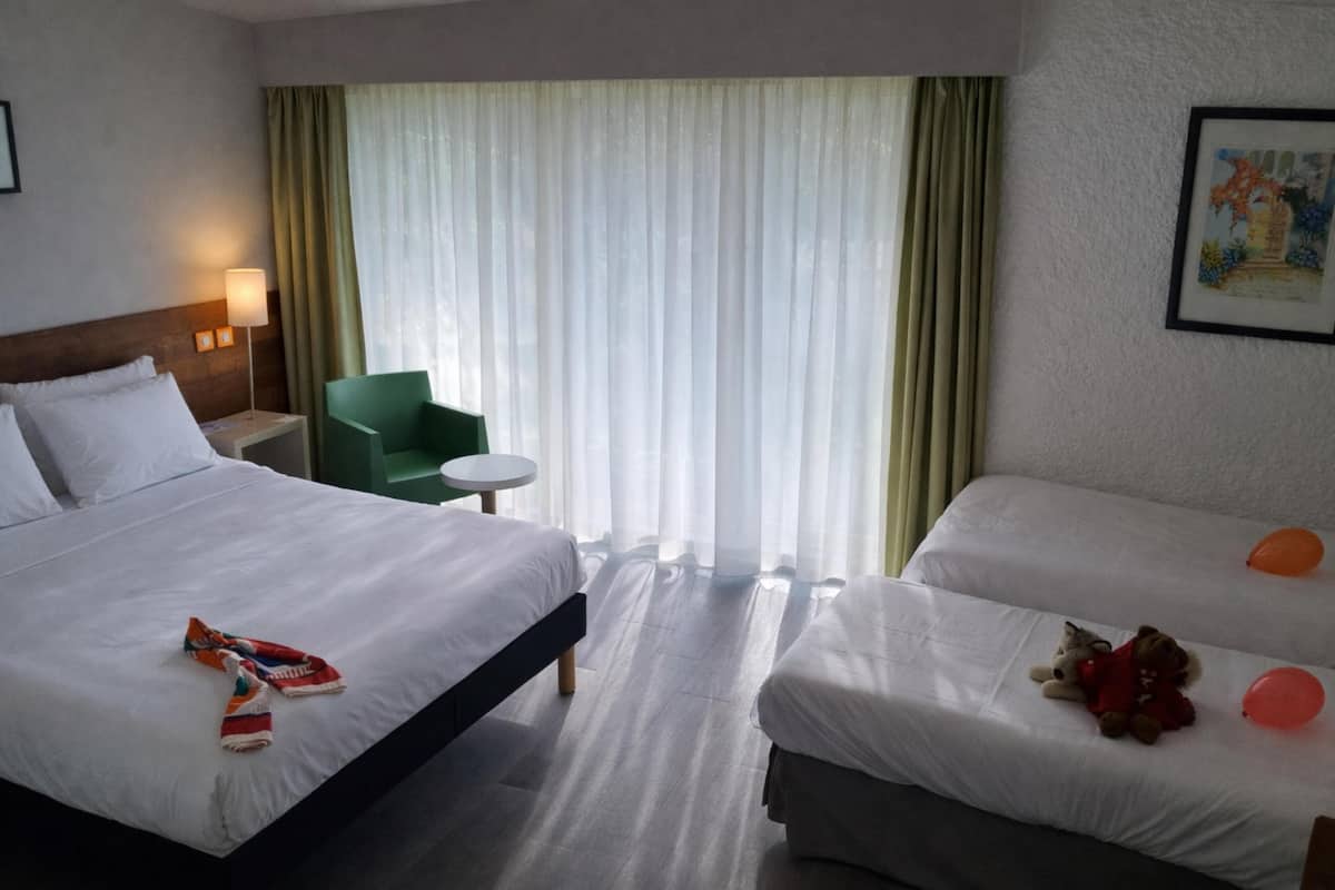Et hotelværelse med to senge, en grøn stol, et rundt bord og et vindue med lette gardiner.
