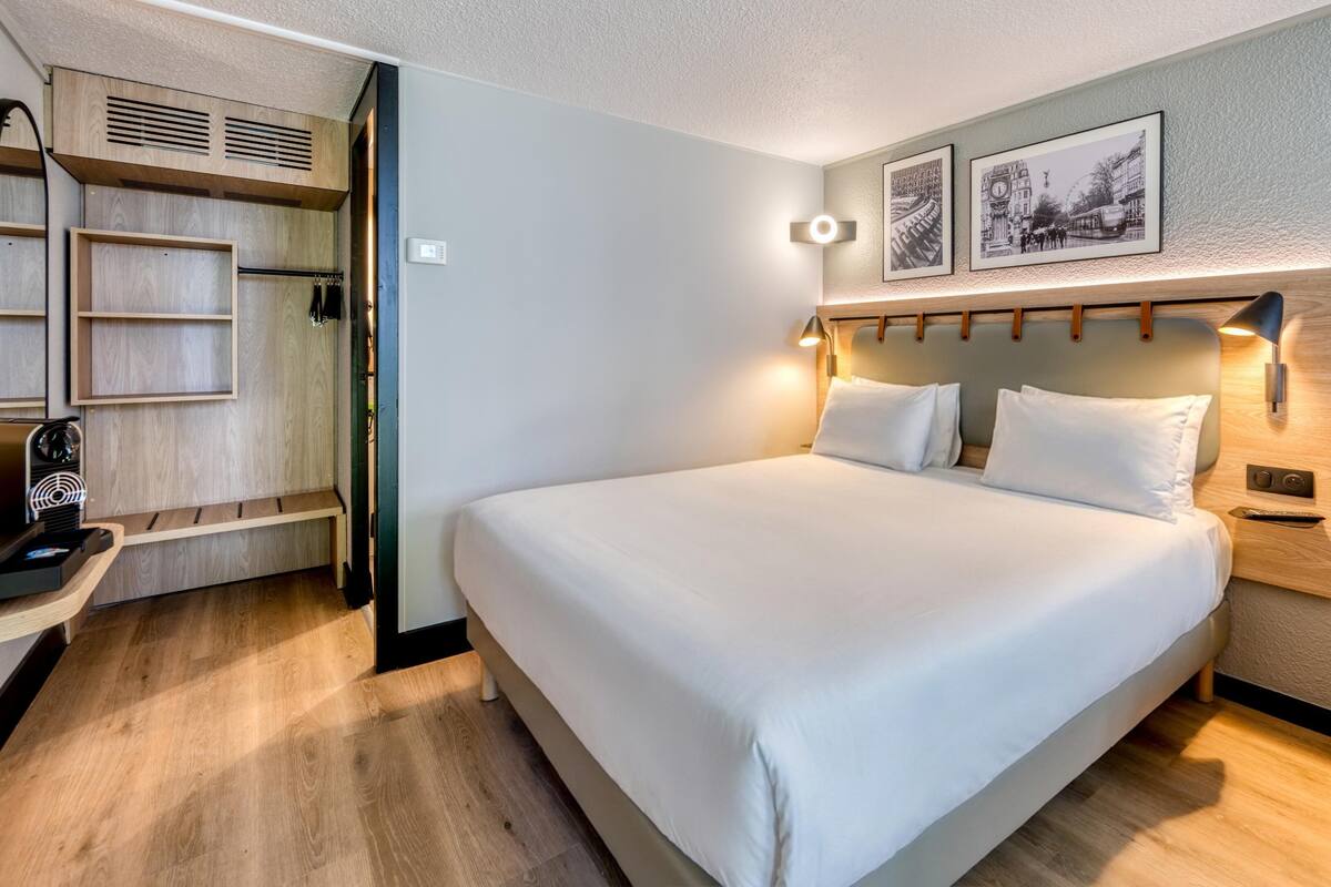 Une chambre d’hôtel avec un grand lit, un bureau avec une machine à café, une armoire et deux tableaux encadrés au mur.