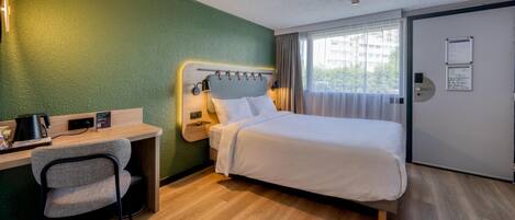 Habitación de hotel con cama, escritorio, silla y vistas a edificios a través del ventanal.