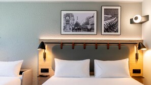 Habitación de hotel con cama, dos almohadas, cabecero con luces fijadas a la pared y dos cuadros enmarcados.