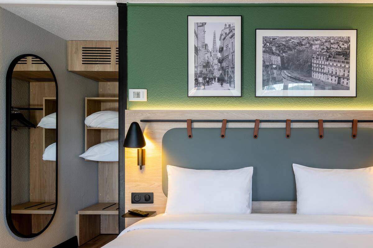 Une chambre d’hôtel avec un lit, deux lampes de chevet, un miroir, une tête de lit et deux tableaux encadrés accrochés au mur.