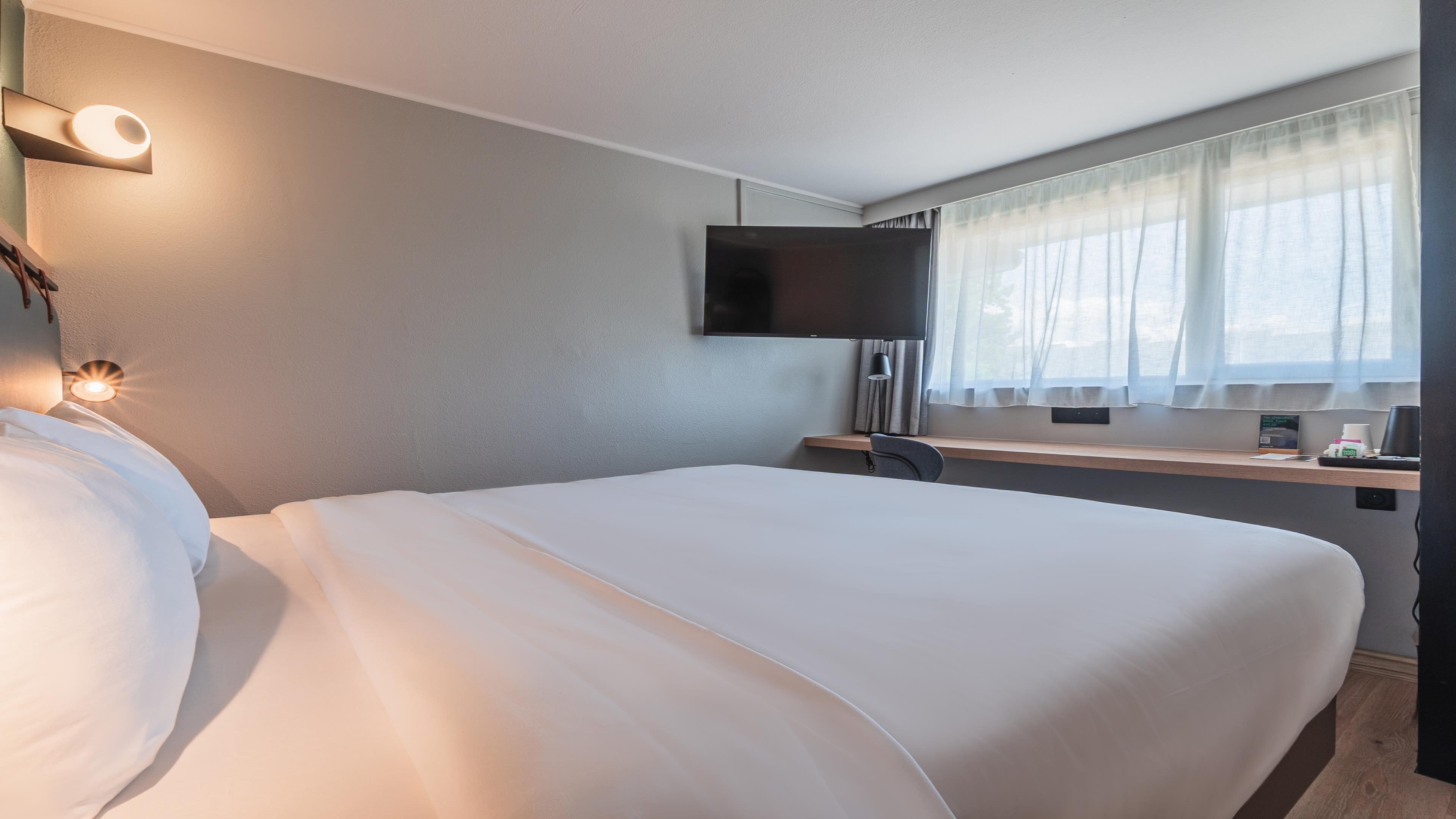 Een hotelkamer met een groot bed, een flatscreen televisie aan de wand, een bureau met stoel, en een raam met dunne gordijnen.
