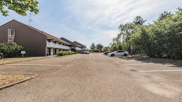 Un parking bordé d’une rangée de bâtiments, avec une voiture et des arbres.