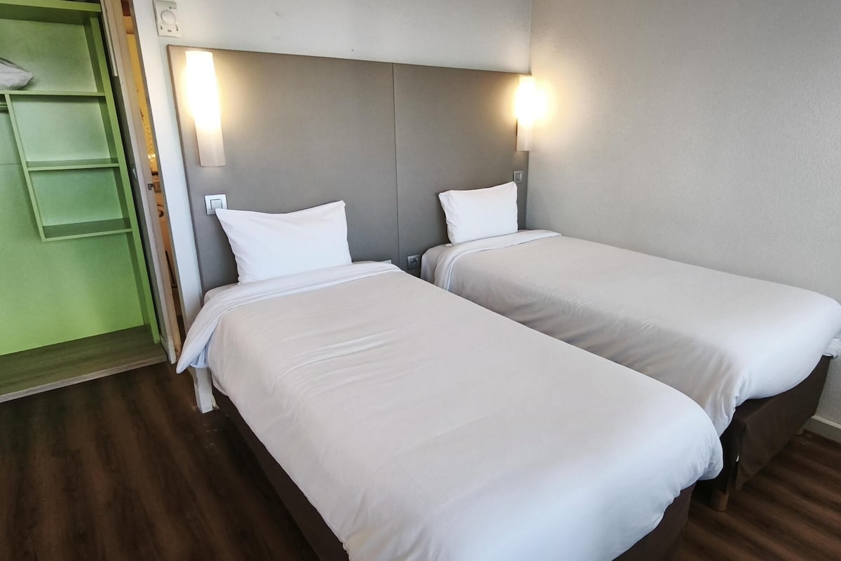 A hotel room with two single beds, a green wardrobe, and a wall-mounted light fixture.