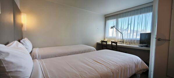 A hotel room with two beds, a desk with a chair, a lamp, a television, and a window with curtains. - Hotel Campanile Beziers (Villeneuve-les-Beziers)