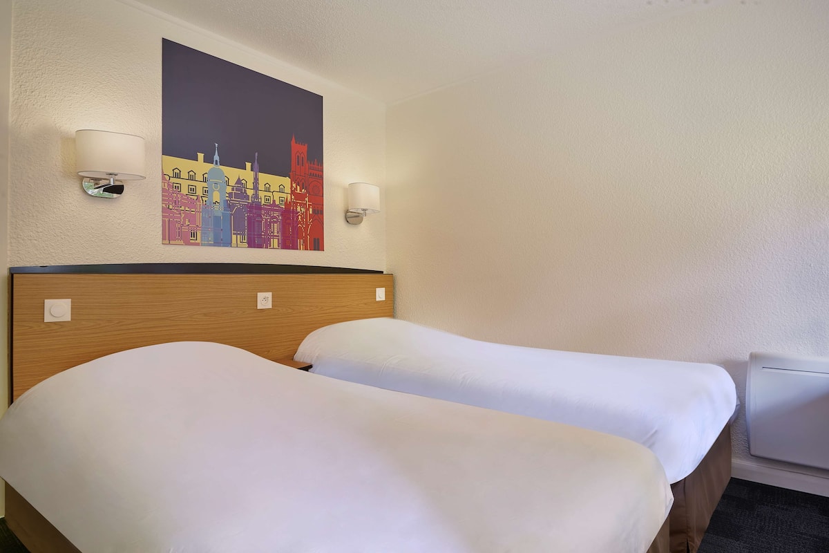A hotel room with two beds, a wooden headboard, two wall-mounted lamps, and a colorful cityscape painting above the beds.