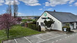 Un bâtiment à l’extérieur blanc, orné d’un logo vert, entouré d’un parking et de verdure.