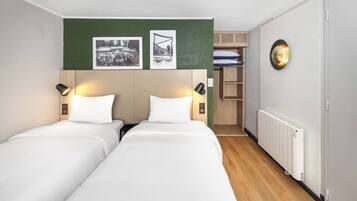 Een hotelkamer met twee enkele bedden, een groene accentmuur, een radiator en een klein schap met opgevouwen handdoeken.