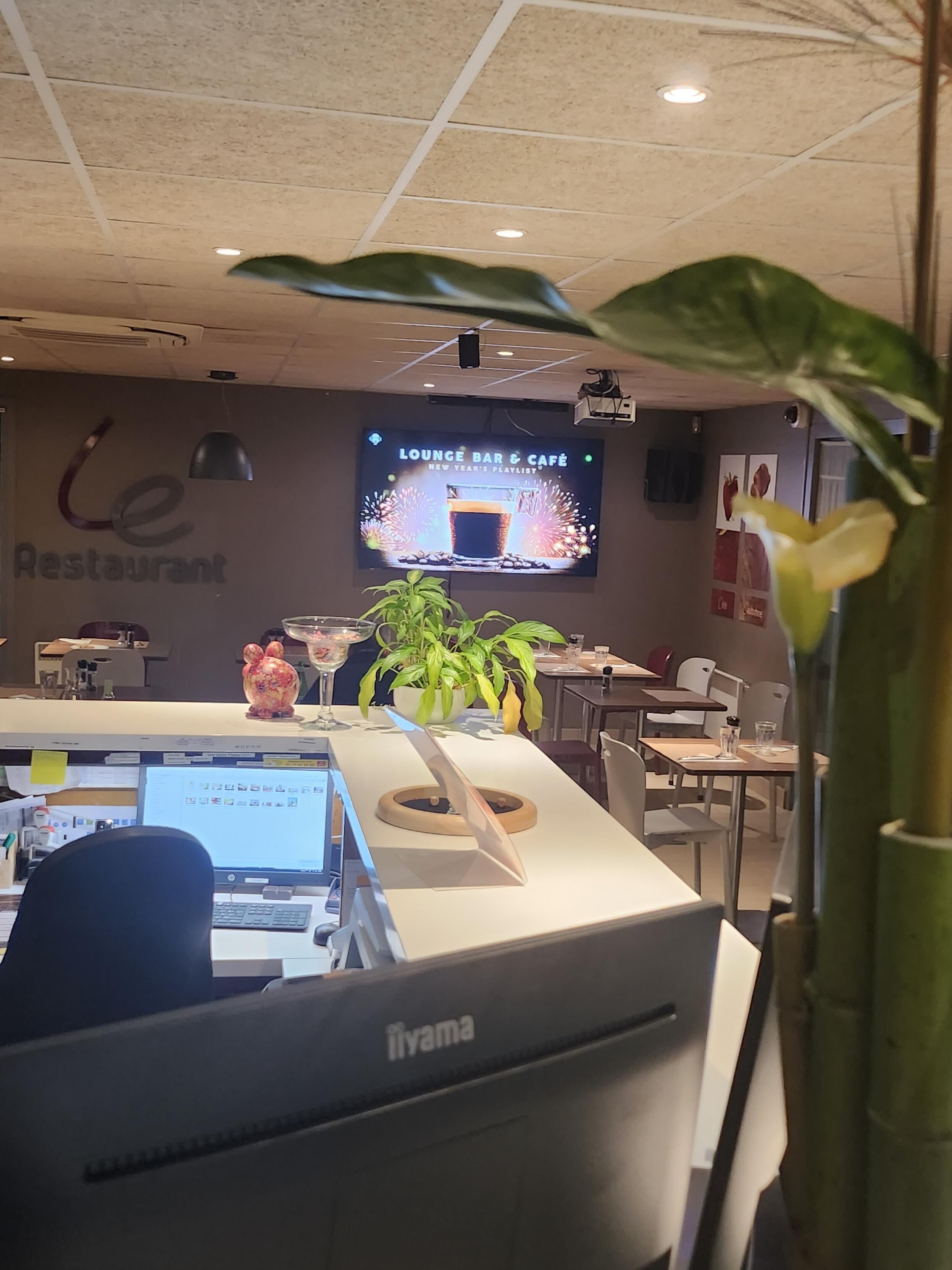 A restaurant with a reception desk, computers, and a TV screen displaying a lounge bar and cafe playlist.