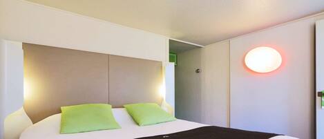 A hotel room with a bed, green pillows, a black throw, and a wall-mounted light.