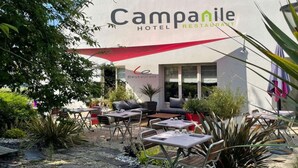 Un área de estar al aire libre con mesas y sillas, un paraguas rojo y un edificio con el letrero "Campanile Hotel Restaurant".