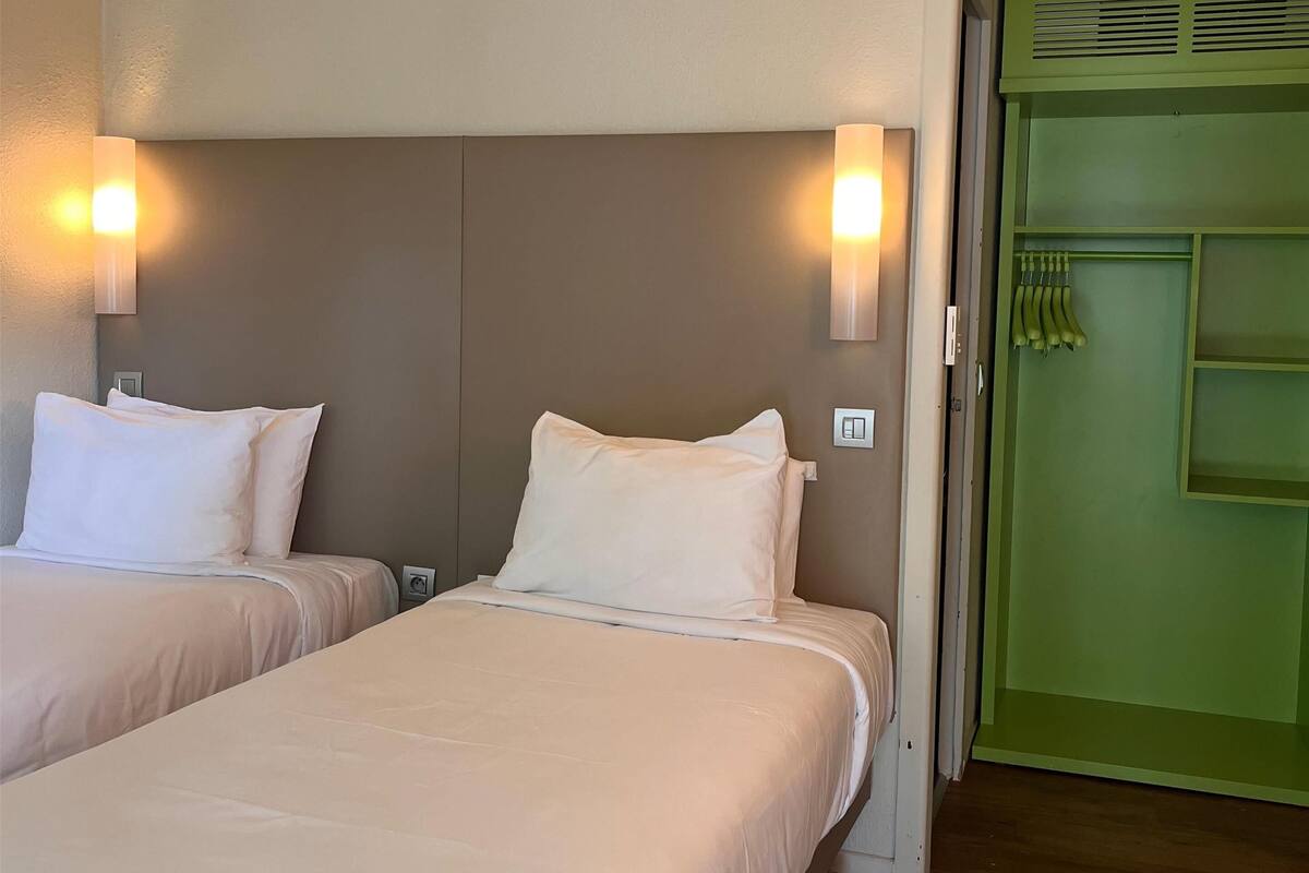A hotel room with two beds, a green door, and a light switch on the wall.